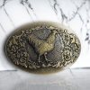 Vintage bronze oval belt buckle with a 3D rooster design, turquoise center stone, and rustic Western finish.