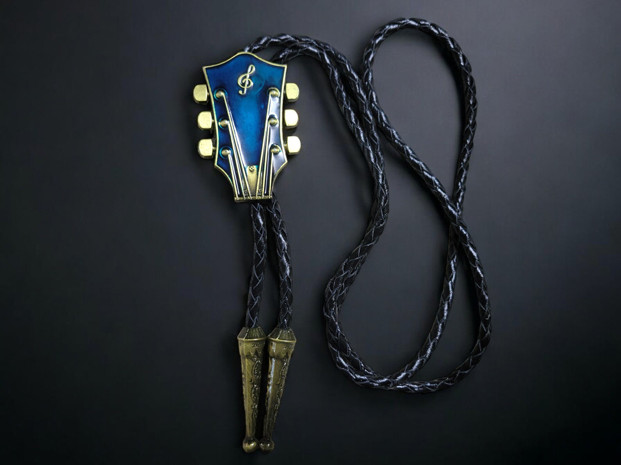 Guitar-shaped bolo tie with braided cowhide leather cord, metal tips, and adjustable slider clasp.